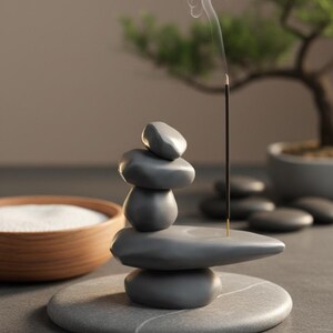 May include: A gray stone incense holder with a stack of smooth, rounded stones. An incense stick is lit, with smoke rising. A wooden bowl with white sand and a bonsai tree are in the background. The holder sits on a round stone base.