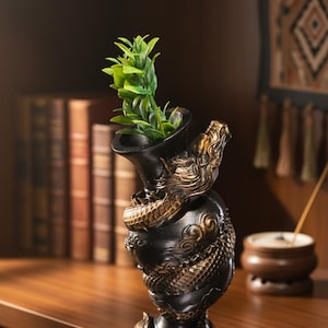 Peut inclure: Vase noir et or en forme de dragon, contenant une plante verte. Le vase est orné d'un dragon détaillé. Il est posé sur une surface en bois, avec des livres et une tenture murale en arrière-plan.