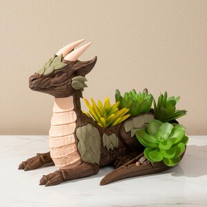 Peut inclure: Un pot en forme de dragon marron avec des détails texturés. Il présente des cornes beiges, une poitrine rose clair et des feuilles vertes. Le pot est rempli de succulentes vertes et jaunes, créant un élément décoratif unique.