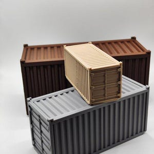 Può includere: Tre container di spedizione in miniatura nei toni del marrone, beige e grigio. I container hanno i lati a coste e sono impilati. Probabilmente vengono utilizzati per il modellismo o l'esposizione.