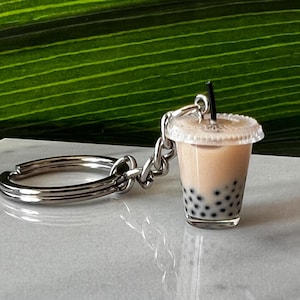 May include: A silver keychain with a clear plastic cup filled with a brown liquid and black tapioca pearls. The cup has a black straw sticking out of it.