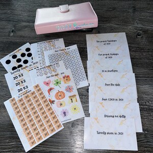 May include: A pale pink savings binder with the text "2021 Savings" and various inserts. The inserts include stickers, calendars, and cards with phrases such as "Save the date" and "Twenty seven in 2021".