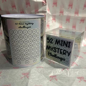 Könnte beinhalten: Zwei Behälter für ein Spiel. Einer ist eine zylindrische Dose mit der Aufschrift "52 Mini-Mystery-Herausforderungen" und nummerierten Kreisen. Der andere ist ein klarer Acryl-Würfel mit demselben Text. Beide stehen auf einer gemusterten Oberfläche.