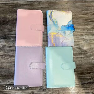 Könnte beinhalten: Vier geschlossene Ordner in Pastellfarben mit Verschluss. Die Ordner sind in Rosa, Lavendel und Hellblau gehalten. Ein Ordner hat ein Marmormuster mit blauen und goldenen Akzenten.