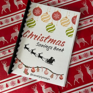 May include: A Christmas Savings Book with a white cover, spiral-bound. The cover displays Christmas baubles and the words "Christmas Savings Book." A silhouette of Santa's sleigh and reindeer is also present, along with the Instagram handle @Lovelylynseys.