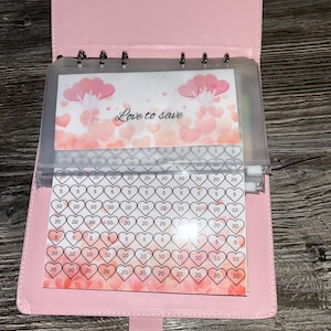 May include: A pink budget binder with clear pockets. The binder has a tab closure and contains inserts with heart-shaped graphics and the text "Love to save." The inserts have spaces for tracking savings goals.