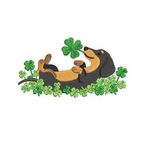 May include: An embroidered design of a dachshund dog lying on its back, surrounded by green shamrocks. The dog is black and tan, with a shamrock in its mouth. The design is on a white background.