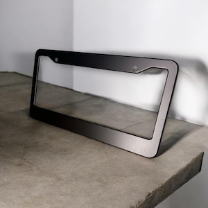May include: A matte black license plate frame with a rectangular shape and rounded corners. The frame has two small circular holes for mounting and is displayed on a concrete surface.