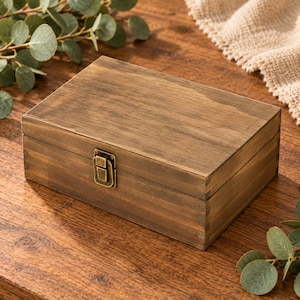 May include: A rectangular wooden storage box with a dark brown stained finish and visible wood grain. The box features a hinged lid and a small, centered antique brass-colored metal latch on the front for secure closure.