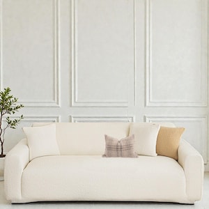 May include: A cream-coloured, modern sofa with a textured surface. The sofa has a rounded shape and is adorned with four decorative cushions in cream, tan, and plaid patterns. A small potted plant is visible on the left side of the image.