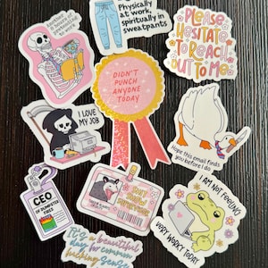 May include: A collection of humorous stickers with various designs and text. The stickers feature phrases like "Didn't punch anyone today," "I love my job," and "Very worky today." Other designs include a skeleton, a grim reaper, and a frog.