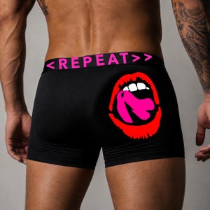 May include: Black boxer briefs featuring a waistband with the pink text <REPEAT>>. The back of the garment displays a large graphic of red lips with white teeth and a bright pink tongue, positioned on the right side of the black fabric.