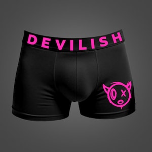 May include: Black boxer briefs featuring a bright pink elastic waistband with the text DEVILISH printed in bold capital letters. A matching bright pink graphic of a stylized devilish character face with horns is positioned on the lower right leg.