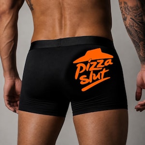 Op de afbeelding: Zwarte boxershort met een brede elastische tailleband en een feloranje afbeelding aan de rechterkant. De afbeelding toont een gestileerd, dakvormig pizza-icoon boven de tekst "pizza slut" in een vet, informeel, kleine lettertype.