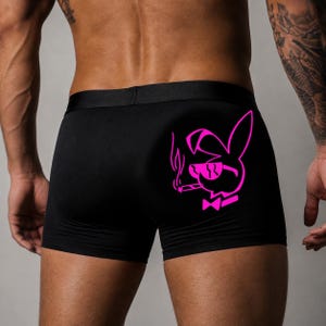 May include: Black boxer briefs featuring a vibrant neon pink graphic on the right side. The design depicts a stylized rabbit silhouette wearing sunglasses and a bow tie, while holding a smoking cigarette. The garment has a wide, solid black waistband.