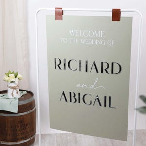May include: A sage green wedding welcome sign with the names "RICHARD and ABIGAIL" in a stylish font. The sign is suspended from a white metal frame with brown leather straps. A small bouquet of white flowers sits on a wooden barrel.