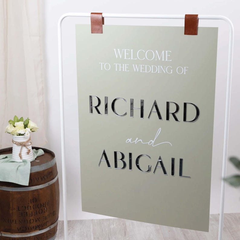 May include: A sage green wedding welcome sign with the names "RICHARD and ABIGAIL" in a stylish font. The sign is suspended from a white metal frame with brown leather straps. A small bouquet of white flowers sits on a wooden barrel.
