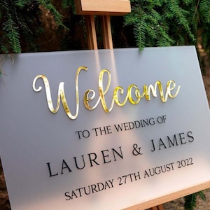 May include: A wedding welcome sign with gold cursive "Welcome" lettering. Below, the sign reads "TO THE WEDDING OF LAUREN & JAMES SATURDAY 27TH AUGUST 2022". The sign is displayed on a wooden easel.