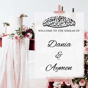 May include: A white sign on a wooden easel with the text "WELCOME TO THE NIKKAH OF Dania & Aymen" in elegant script. The sign is adorned with Arabic calligraphy and floral arrangements, suggesting a wedding or celebration.