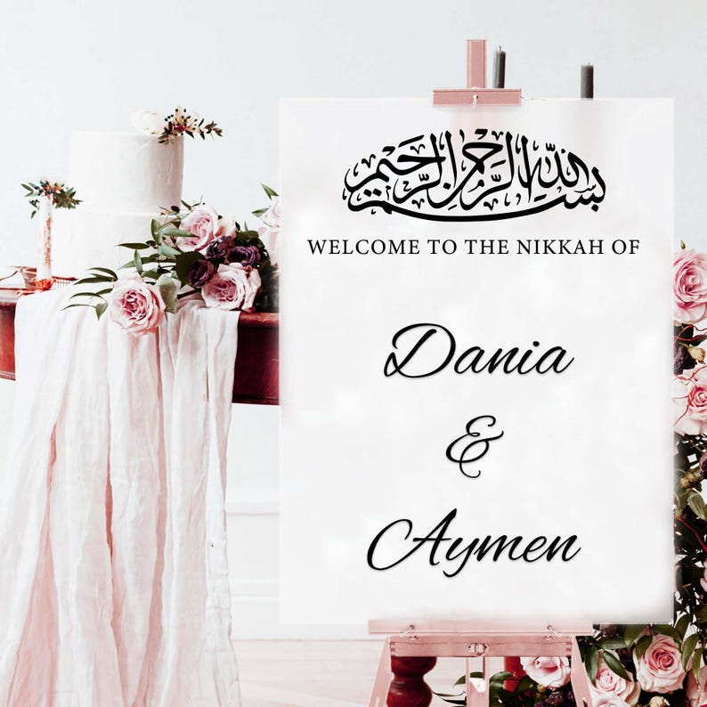 May include: A white sign on a wooden easel with the text "WELCOME TO THE NIKKAH OF Dania & Aymen" in elegant script. The sign is adorned with Arabic calligraphy and floral arrangements, suggesting a wedding or celebration.