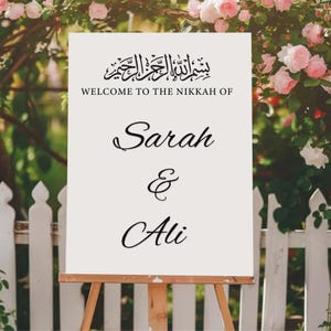 May include: A white sign on a wooden easel, featuring the text "WELCOME TO THE NIKKAH OF" above the names "Sarah & Ali" in cursive script. The sign is set against a backdrop of a white picket fence and pink roses.