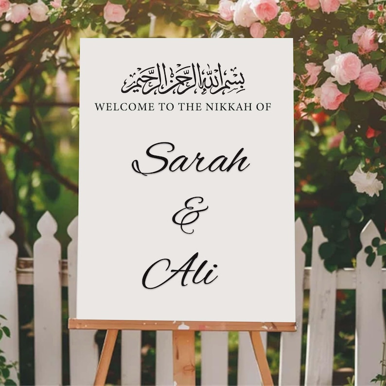 May include: A white sign on a wooden easel, featuring the text "WELCOME TO THE NIKKAH OF" above the names "Sarah & Ali" in cursive script. The sign is set against a backdrop of a white picket fence and pink roses.