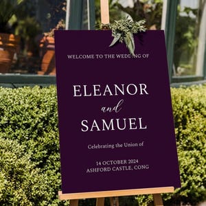 May include: A dark purple wedding welcome sign on an easel. The sign reads "WELCOME TO THE WEDDING OF ELEANOR and SAMUEL" with the date and location below. Greenery is placed at the top of the sign.
