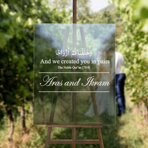 May include: A clear acrylic wedding sign displayed on a wooden easel. The sign has Arabic script and English text: "And we created you in pairs" and the names "Aras and Ikram". The background is a blurred outdoor scene.