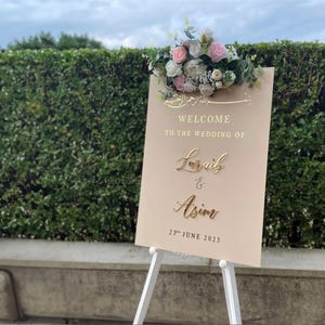 May include: A wedding welcome sign on a white easel. The sign is a light beige colour with gold lettering that reads "Welcome to the wedding of Loraib & Asim 23rd June 2023." The sign is topped with a floral arrangement of pink and white roses.