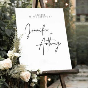 May include: A white wedding sign on a wooden easel, with the text "Welcome to the Wedding of Jennifer + Anthony" in black script. The sign is decorated with white roses and greenery at the bottom.