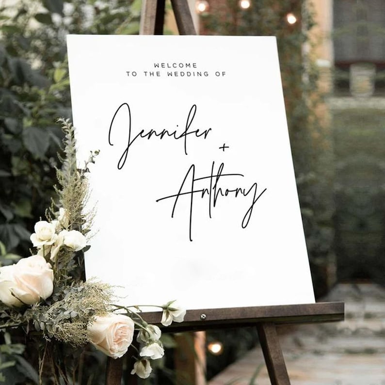May include: A white wedding sign on a wooden easel, with the text "Welcome to the Wedding of Jennifer + Anthony" in black script. The sign is decorated with white roses and greenery at the bottom.