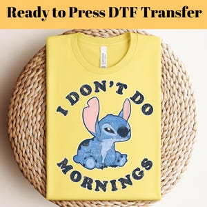 Puede incluir: Camiseta amarilla con el texto "I DON'T DO MORNINGS" y un personaje de dibujos animados azul. La camiseta est&aacute; doblada sobre una superficie tejida. Las palabras "Ready to Press DTF Transfer" est&aacute;n en la parte superior.
