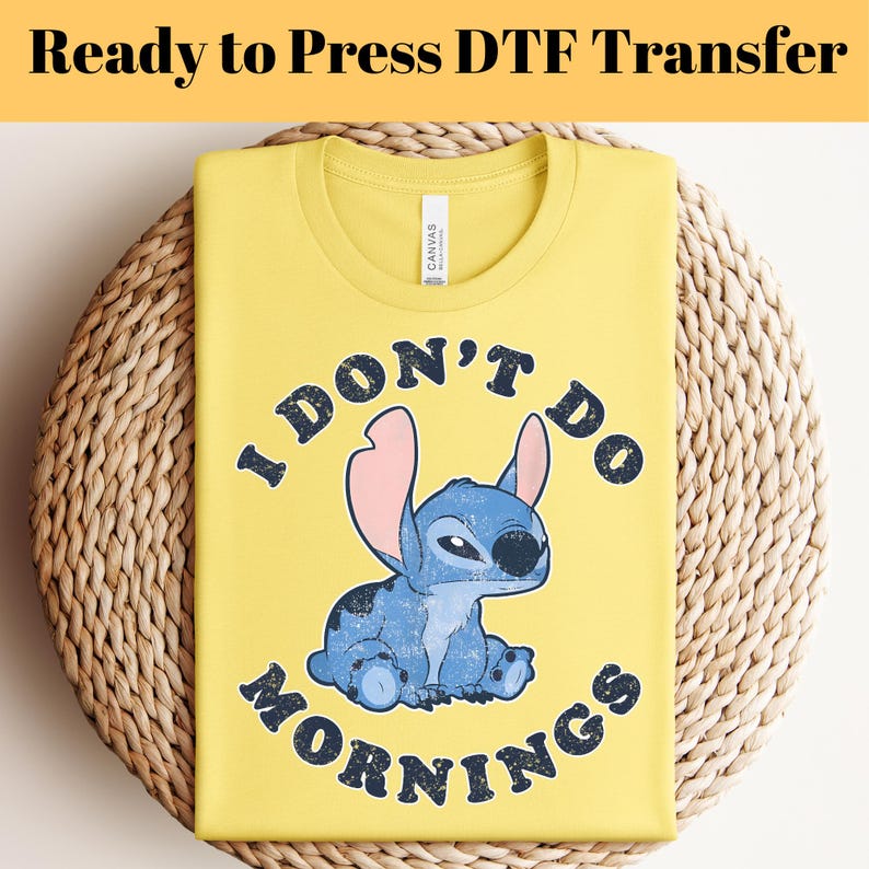 Puede incluir: Camiseta amarilla con el texto "I DON'T DO MORNINGS" y un personaje de dibujos animados azul. La camiseta est&aacute; doblada sobre una superficie tejida. Las palabras "Ready to Press DTF Transfer" est&aacute;n en la parte superior.