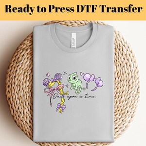 Könnte beinhalten: Ein hellgraues T-Shirt mit einer bunten Grafik mit einem grünen Chamäleon, einer rosa Schleife und dem Text "Once upon a time". Das T-Shirt ist auf einer gewebten Oberfläche gefaltet, mit dem Text "Ready to Press DTF Transfer" oben.