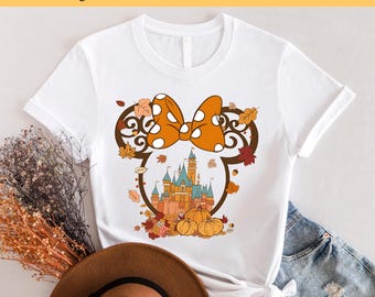 May include: White t-shirt with a  fall-themed design featuring a Mickey Mouse silhouette with a bow and Cinderella's castle inside. The design includes orange, brown, and white colors.