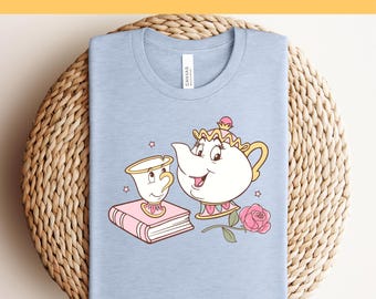 Könnte beinhalten: Hellblaues T-Shirt mit einem Aufdruck einer Teekanne und einer Teetasse aus dem Disney-Film Die Schöne und das Biest. Die Teekanne ist weiß mit einem rosafarbenen Deckel und einer rosafarbenen Rose an der Seite. Die Teetasse ist gelb mit einem rosafarbenen Henkel und einer rosafarbenen Rose an der Seite. Der Aufdruck ist von rosafarbenen Sternen umgeben.