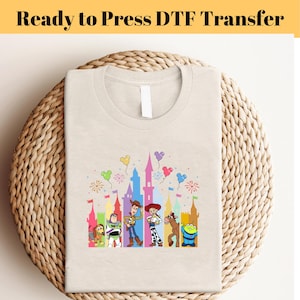 Può includere: Una maglietta beige con un design di trasferimento DTF che presenta i personaggi di Toy Story, Woody, Buzz Lightyear, Jessie e altri davanti a una silhouette di castello colorata. Il design include palloncini e fuochi d'artificio.