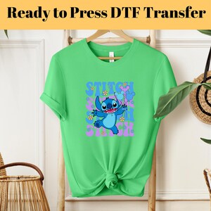 May include: Green t-shirt with a blue Stitch character from Lilo & Stitch. Stitch is holding a purple flower and surrounded by pink flowers. The text "Stitch Ohana Stitch" is printed on the shirt.