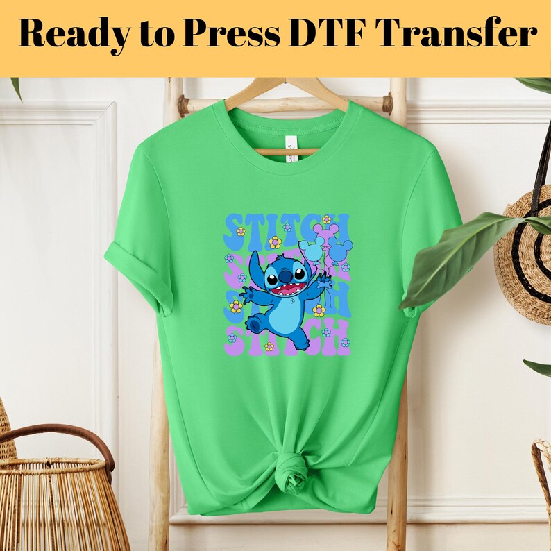 May include: Green t-shirt with a blue Stitch character from Lilo & Stitch. Stitch is holding a purple flower and surrounded by pink flowers. The text "Stitch Ohana Stitch" is printed on the shirt.