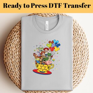 Pode incluir: Uma camiseta cinza com um gr&aacute;fico colorido dos personagens do Toy Story em uma x&iacute;cara de ch&aacute; com bal&otilde;es. Os personagens incluem Woody, Buzz Lightyear, Jessie, Rex e Slinky Dog.