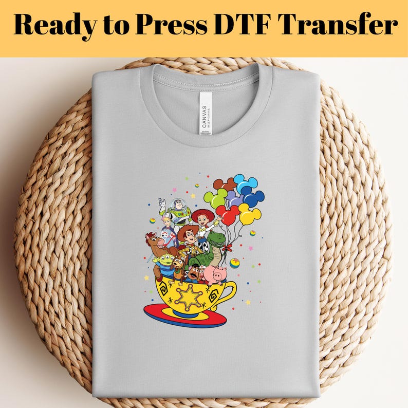 Pode incluir: Uma camiseta cinza com um gr&aacute;fico colorido dos personagens do Toy Story em uma x&iacute;cara de ch&aacute; com bal&otilde;es. Os personagens incluem Woody, Buzz Lightyear, Jessie, Rex e Slinky Dog.