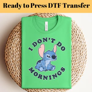 Puede incluir: Una camiseta verde lima con un personaje de dibujos animados azul y el texto "I DON'T DO MORNINGS". La camiseta est&aacute; doblada sobre una superficie circular tejida. Las palabras "Ready to Press DTF Transfer" est&aacute;n en la parte superior.