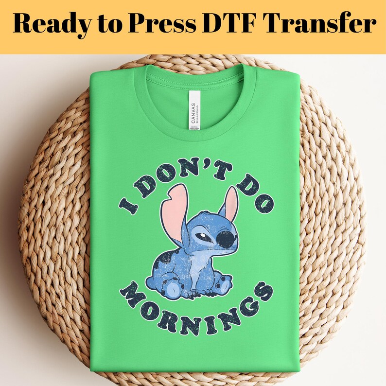 Puede incluir: Una camiseta verde lima con un personaje de dibujos animados azul y el texto "I DON'T DO MORNINGS". La camiseta est&aacute; doblada sobre una superficie circular tejida. Las palabras "Ready to Press DTF Transfer" est&aacute;n en la parte superior.