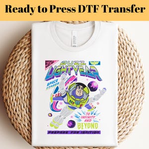 Puede incluir: Camiseta blanca con un gráfico colorido de Buzz Lightyear. El diseño incluye el texto "BUZZ LIGHTYEAR", "TO INFINITY AND BEYOND" y "PREPARE FOR IGNITION". La camiseta está doblada sobre una superficie tejida.