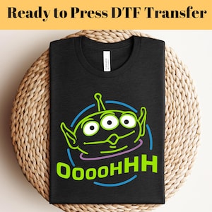 Puede incluir: Una camiseta negra con un personaje extraterrestre verde con tres ojos y el texto "OOOOHHH" en una fuente verde neón y azul.