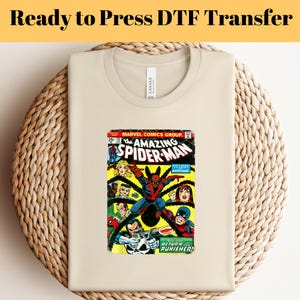 May include: A beige t-shirt with a vintage comic book print featuring Spider-Man and the Punisher. The comic book cover is red, yellow, and blue with the text "The Amazing Spider-Man" and "Return of the Punisher!"