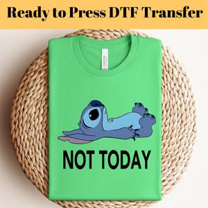 K&ouml;nnte beinhalten: Ein gr&uuml;nes T-Shirt mit einer blauen Cartoon-Figur und dem schwarzen Text "NOT TODAY". Das T-Shirt ist auf einer gewebten Matte gefaltet. Die Worte "Ready to Press DTF Transfer" stehen oben im Bild.