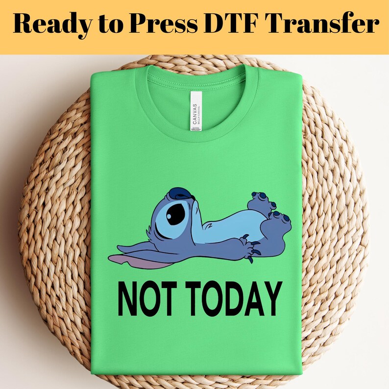 K&ouml;nnte beinhalten: Ein gr&uuml;nes T-Shirt mit einer blauen Cartoon-Figur und dem schwarzen Text "NOT TODAY". Das T-Shirt ist auf einer gewebten Matte gefaltet. Die Worte "Ready to Press DTF Transfer" stehen oben im Bild.