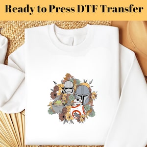 Puede incluir: Sudadera blanca con un gráfico colorido que presenta personajes de Star Wars y detalles florales. El diseño incluye un casco de Stormtrooper, un casco mandaloriano, BB-8 y Grogu, rodeados de flores. El texto "Ready to Press DTF Transfer" está en la parte superior.