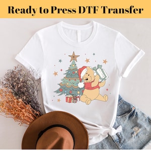 May include: White t-shirt with a Christmas tree and Winnie the Pooh holding a gift. The design is in a vintage style with a red, green, and gold color scheme.  "Ready to Press DTF Transfer" is written in yellow at the top of the image.